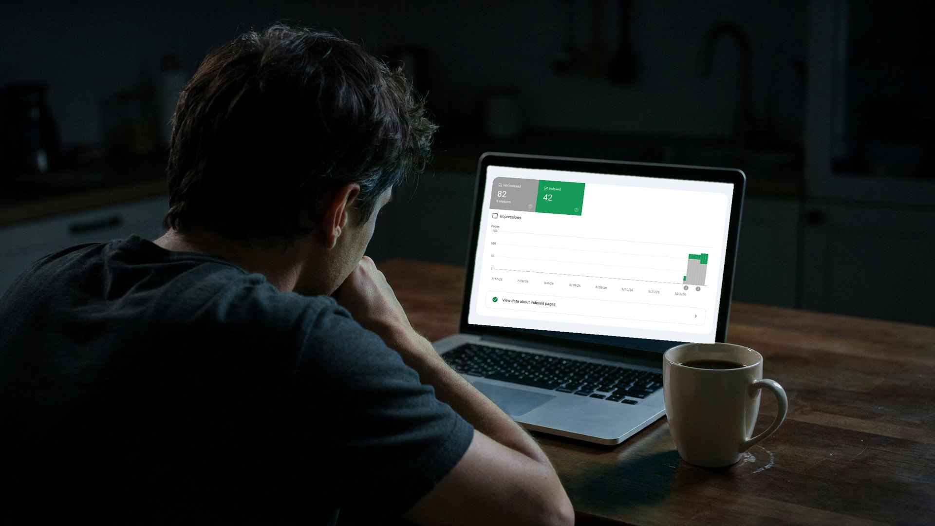 A man waits late at night at his kitchen table, chin on fist, in front of a laptop showing a Search Console indexing chart, his coffee gone cold.
